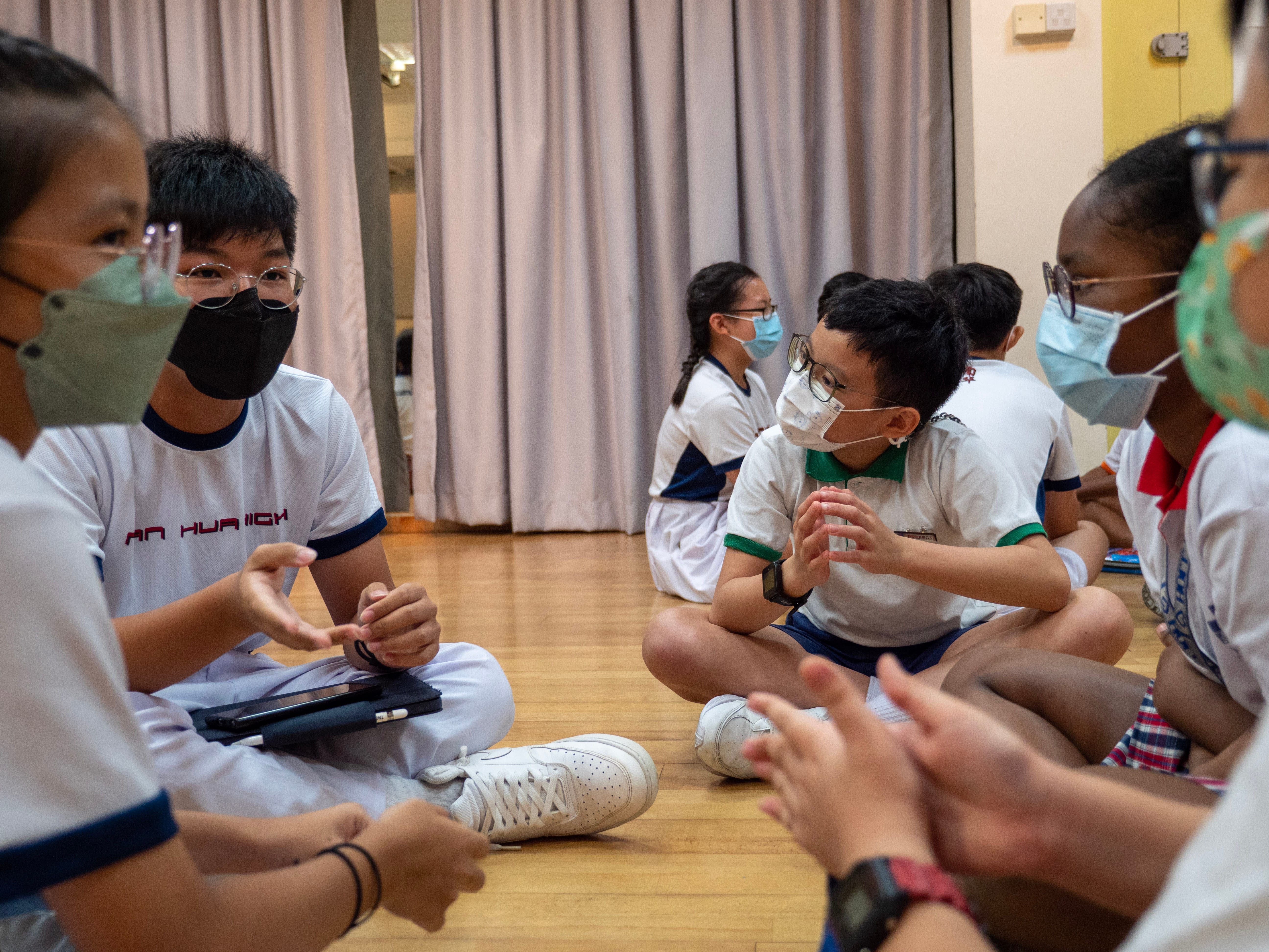 Masked students sit on a gym floor in a circle, talking; curtains hang behind them.